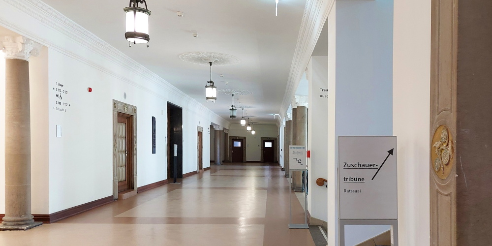 Ein breiter Gang, links im Vordergrund eine Säule, dahinter gepolsterte Türen, rechts vorne ein Schild mit einem Wegweiser zu einer Besuchertribüne oben