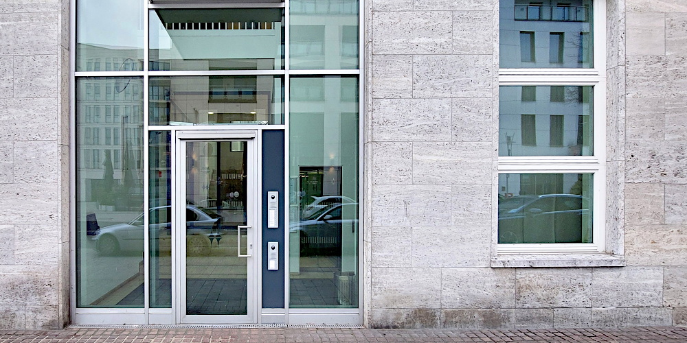 Ausschnitt einer Fassade, links eine hohen Glasfläche mit Tür, rechts ein Fenster